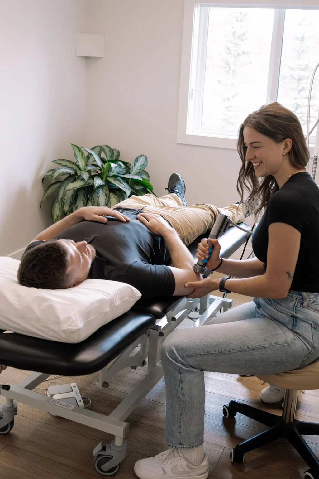 A woman is performing Gunn IMS on a man lying on a medical examination table in a bright room with a window and a plant in the background.