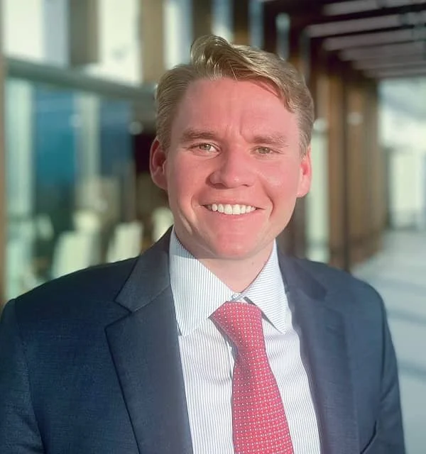 Portrait of a young man in a suit, smiling, in an indoor setting with large windows.