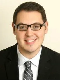 Headshot of a smiling man with glasses, wearing a black suit and tie, against a plain light-colored background.