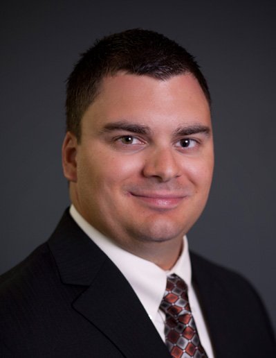 Professional headshot of a man in a black suit, white shirt, and patterned tie, smiling against a dark gray background.