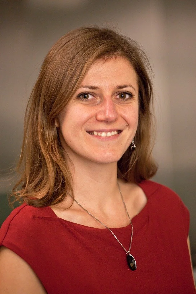 A woman with shoulder-length light brown hair, wearing a red top, earrings, and a necklace with a black pendant, smiling at the camera.
