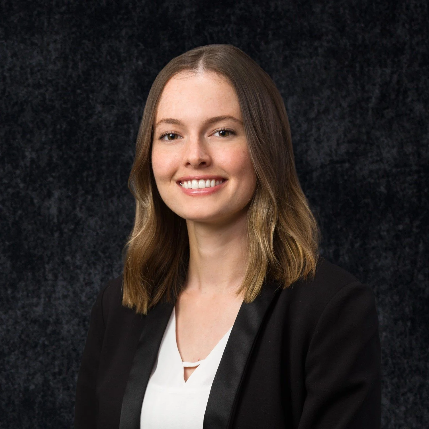 A portrait of a young woman with shoulder-length light brown hair, smiling, wearing a black blazer over a white top, against a dark textured background.