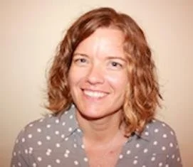 A woman with shoulder-length curly red hair, smiling, wearing a gray shirt with white polka dots, against a plain light-colored background.