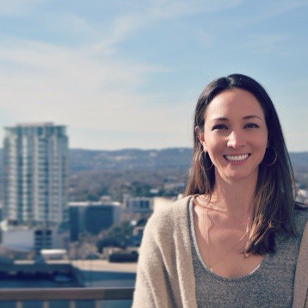 Smiling woman with long dark hair and hoop earrings standing outdoors with city skyline and blue sky in the background.