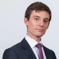 Portrait of a young man in a dark suit with a white shirt and purple tie, against a plain light background.