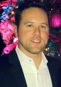 A man dressed in a white shirt and black blazer standing in front of a decorated colorful Christmas tree.