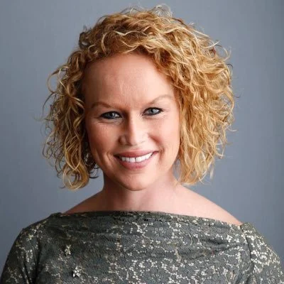 Close-up portrait of a woman with curly blonde hair smiling at the camera against a gray background.