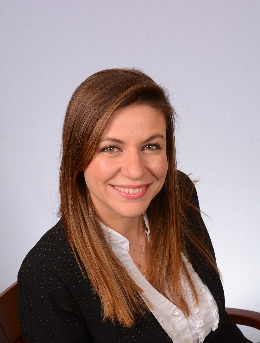 Professional woman with long brown hair, wearing a black blazer and white blouse, smiling at the camera against a plain light background.