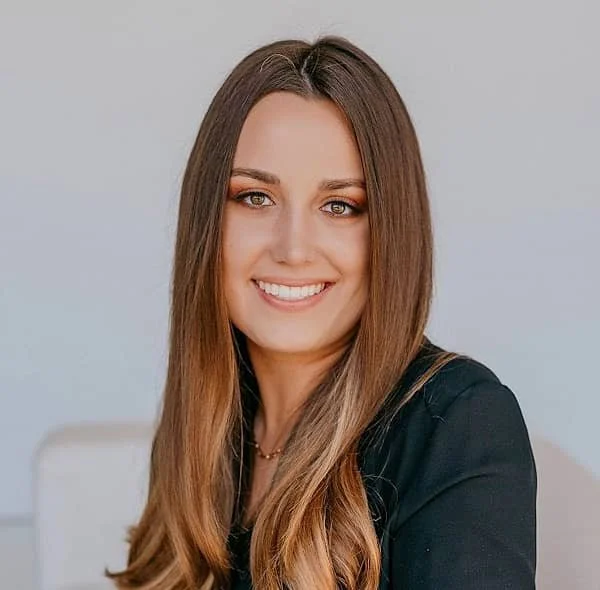 A woman with long light brown hair, smiling, wearing a black top, against a plain light background.