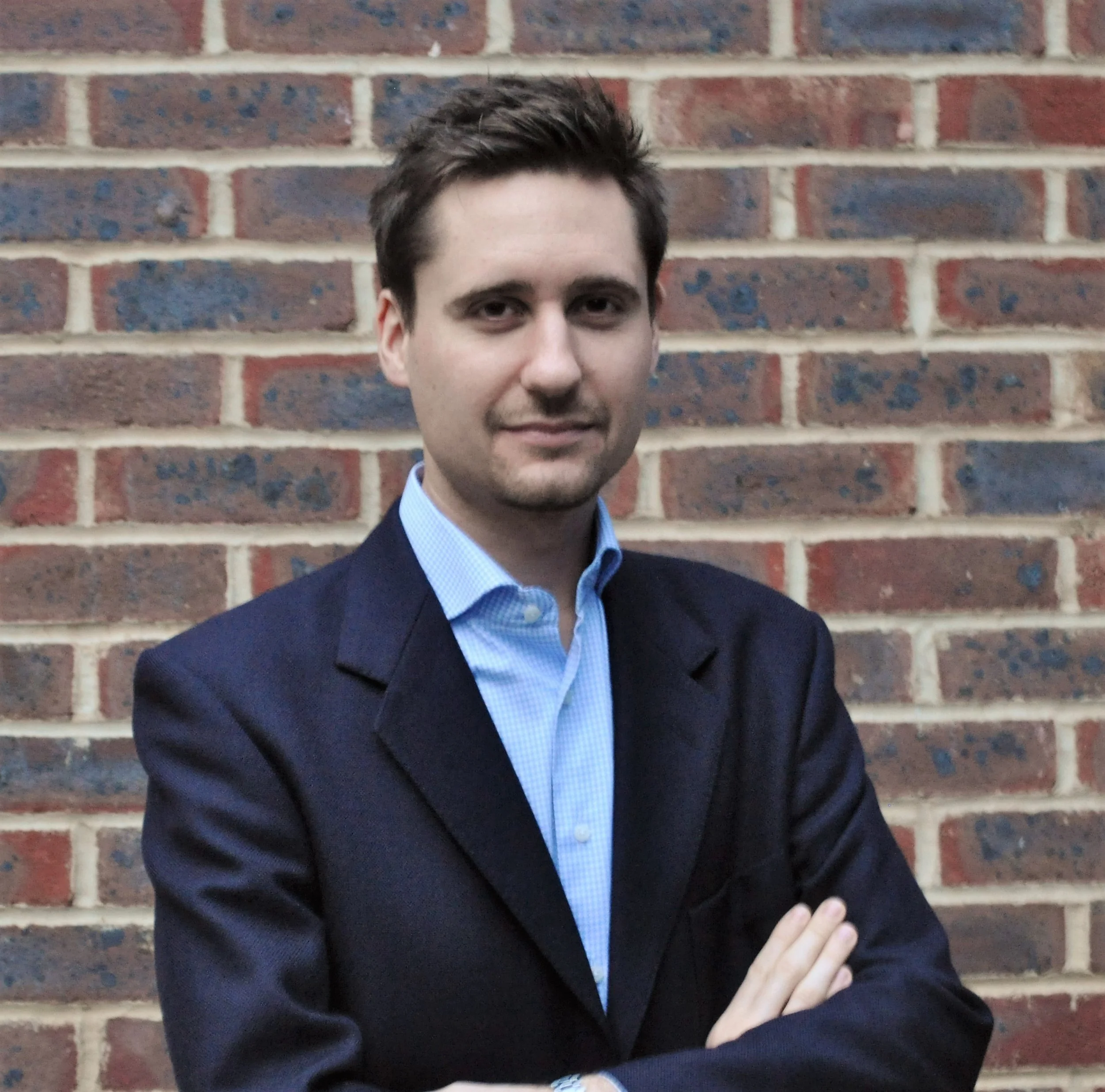 A man with brown hair, dressed in a dark blazer and light blue shirt, standing with arms crossed in front of a brick wall.