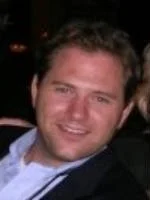 A young man with short curly hair smiling at the camera, wearing a black jacket and a white shirt.