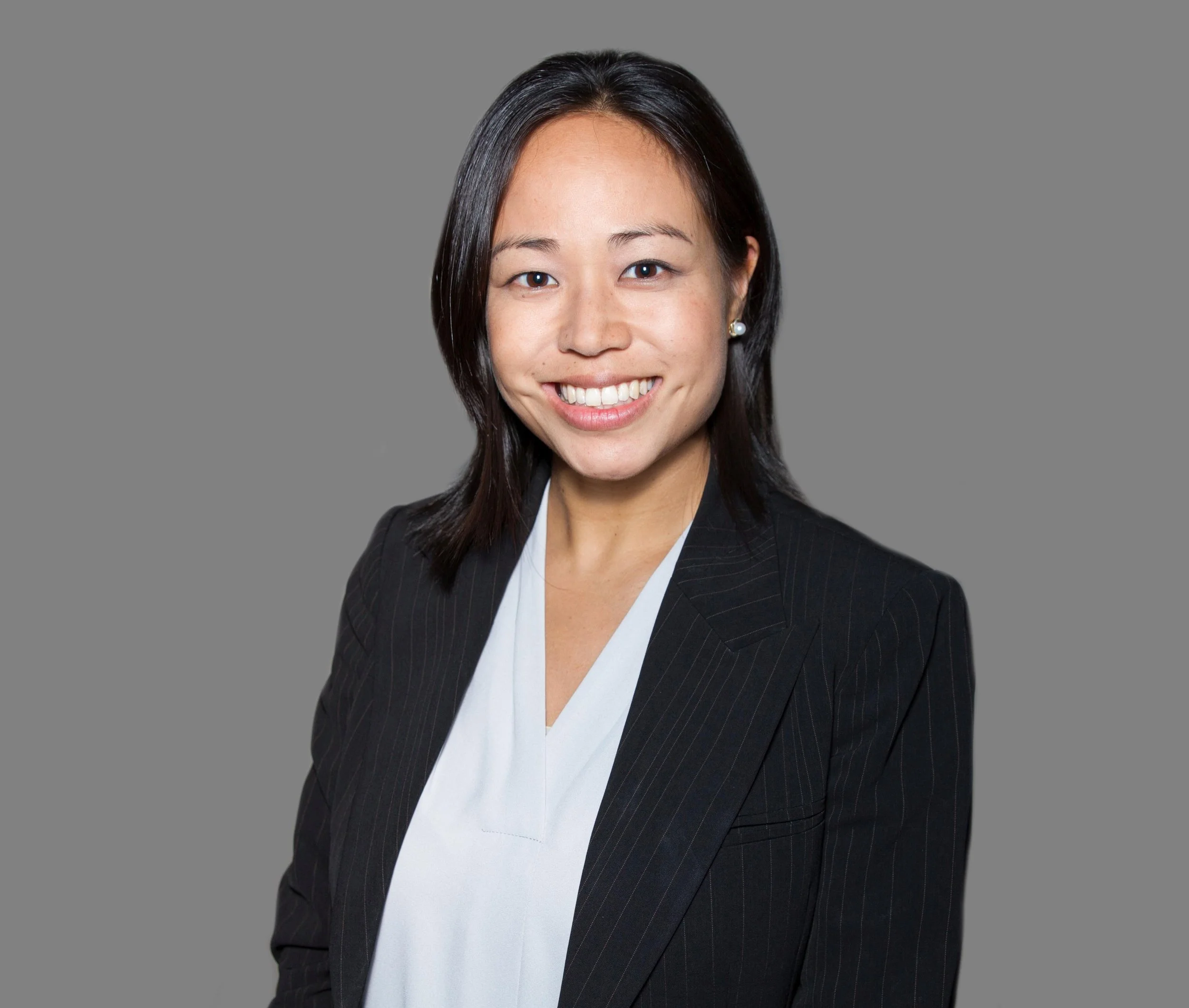 Professional woman with shoulder-length dark hair and pearl earrings, smiling and wearing a black pinstripe blazer and white blouse against a gray background.