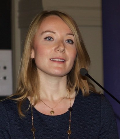 A woman with blonde hair and a dark blue top speaking at a microphone in an indoor setting.
