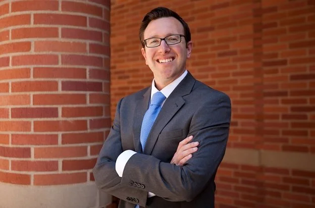 A smiling man in a business suit and glasses standing with arms crossed outside near a brick building.