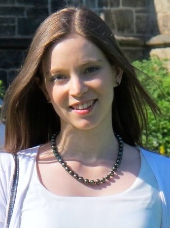 A young woman with long brown hair, smiling, wearing a white top and a black beaded necklace outdoors.