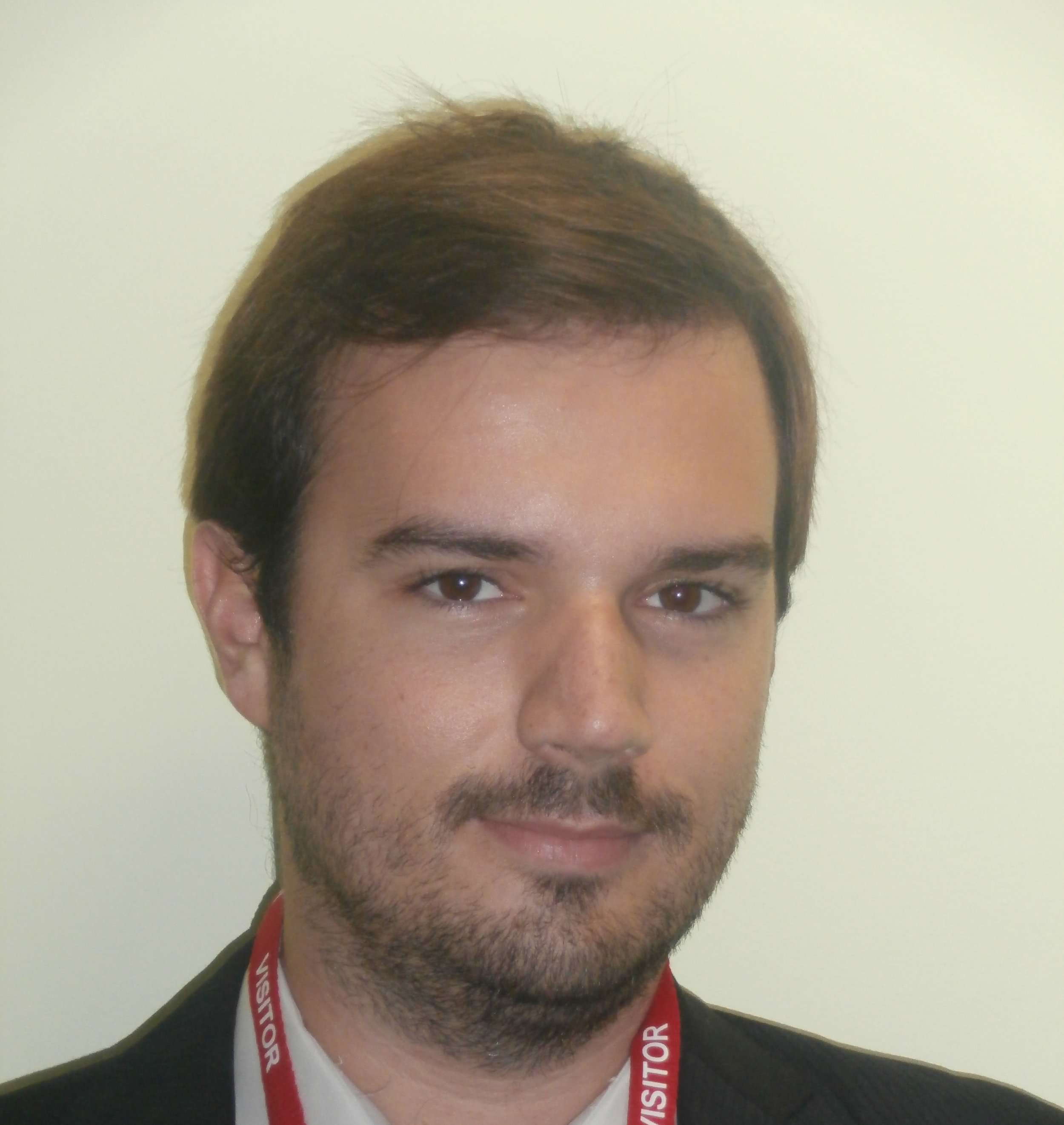 A man with short brown hair and a beard, wearing a black suit and a lanyard that says 'VISITOR'.