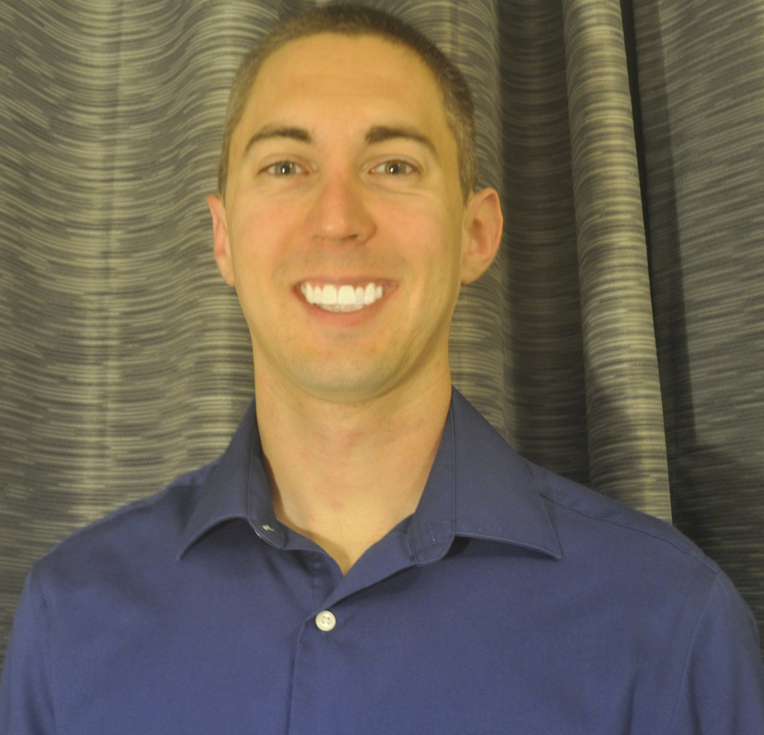 A smiling man wearing a blue collared shirt standing in front of a textured curtain.