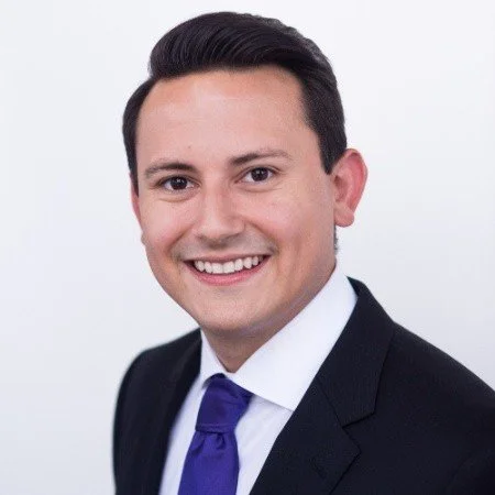 Professional headshot of a man in a black suit, white shirt, and purple tie, smiling against a white background.