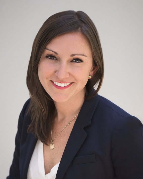 Professional woman with brown hair, smiling, wearing a navy blazer, white top, gold necklace, and earrings, against a light background.