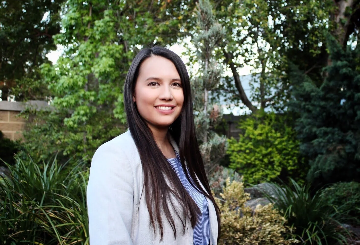 A young woman with long dark hair smiling outdoors in a lush garden with various green bushes and trees.