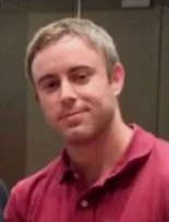 A young man with short hair wearing a maroon polo shirt, standing indoors against a neutral background.