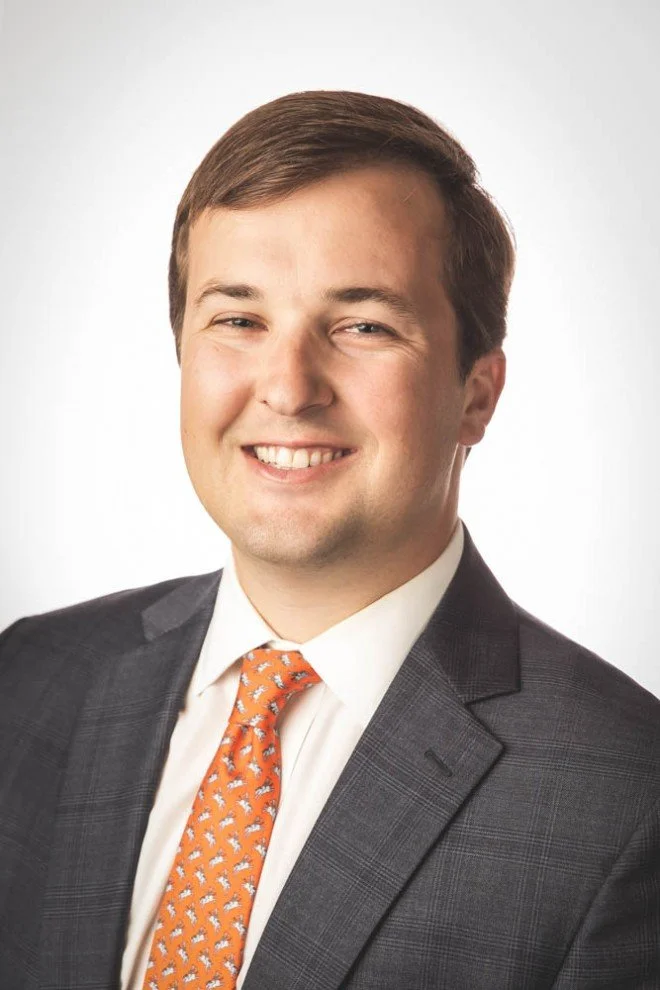 A young man with brown hair in a suit, white shirt, and orange patterned tie, smiling for a professional portrait against a plain white background.