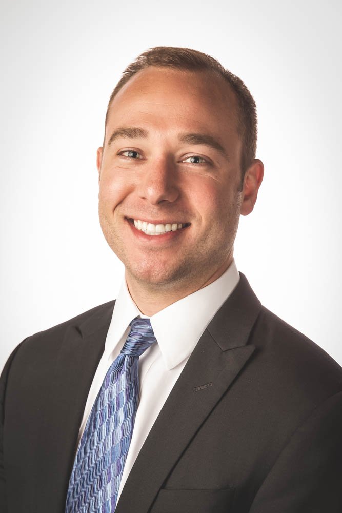 A professional headshot of a smiling man in a dark suit, white shirt, and striped blue tie, against a plain white background.
