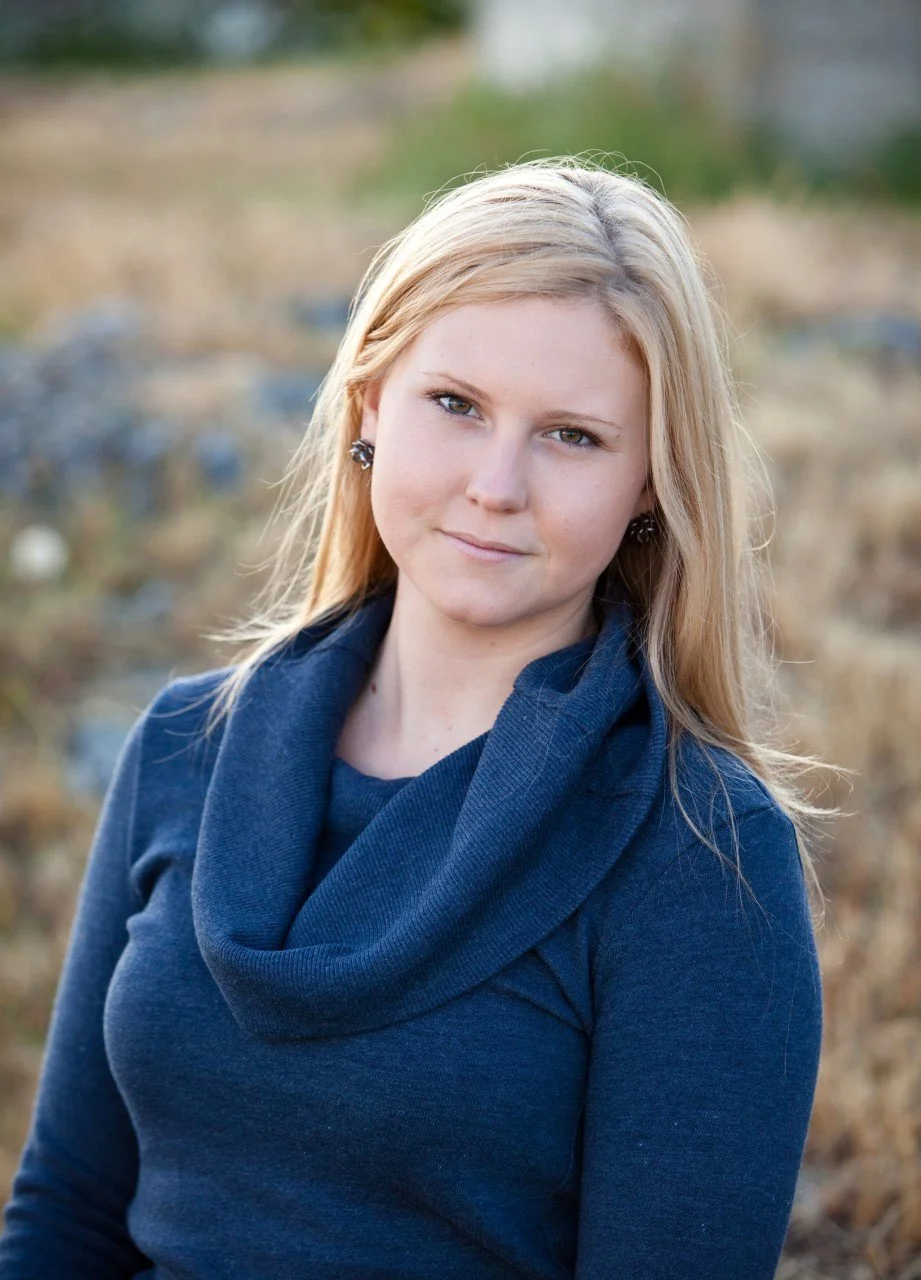 A young woman with blonde hair, blue eyes, and earrings, wearing a blue sweater with a cowl neck, standing outdoors with a blurred natural background.