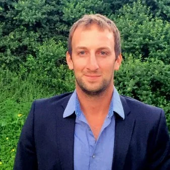 A man with short light brown hair, light facial hair, wearing a blue dress shirt and dark blazer, standing outdoors with green trees and grass in the background.