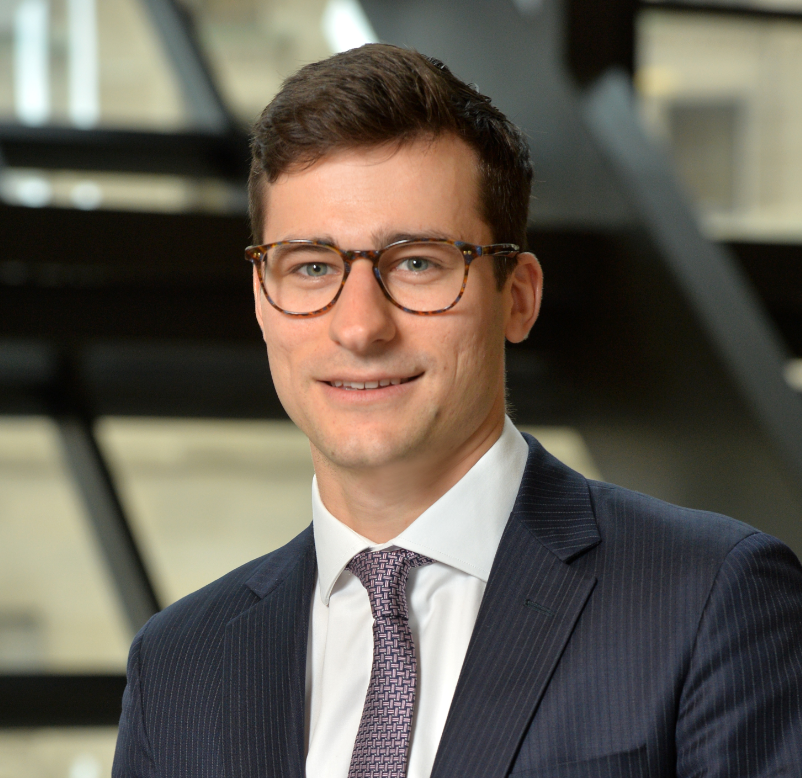 A young man in a dark suit, white shirt, and patterned tie, wearing glasses, smiling in an office building.