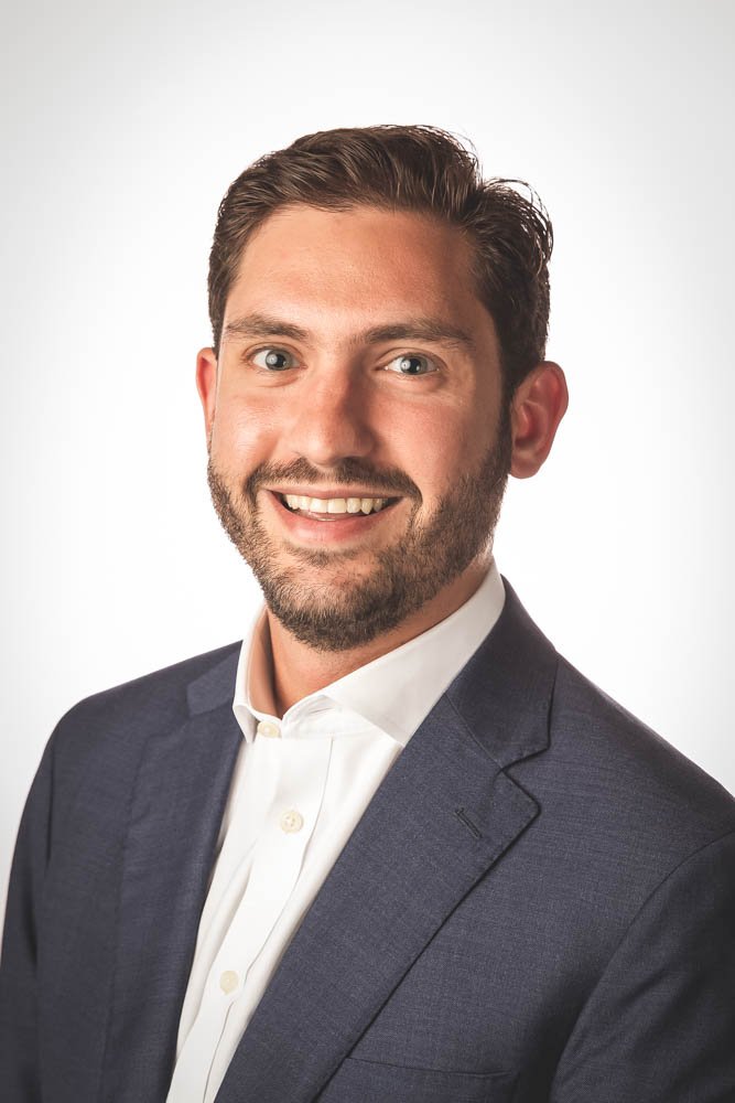 A smiling man with dark hair and a beard, wearing a dark suit jacket and white dress shirt, against a plain white background.