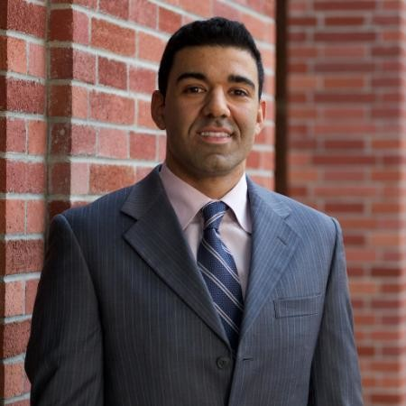 A man in a gray pinstripe suit, light-colored shirt, and striped tie, standing outdoors against a red brick wall, smiling slightly.