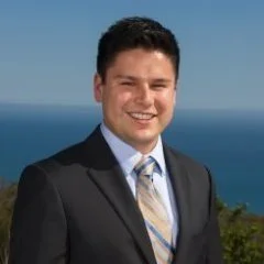 A young man in a suit smiling outdoors with mountains and a body of water in the background.