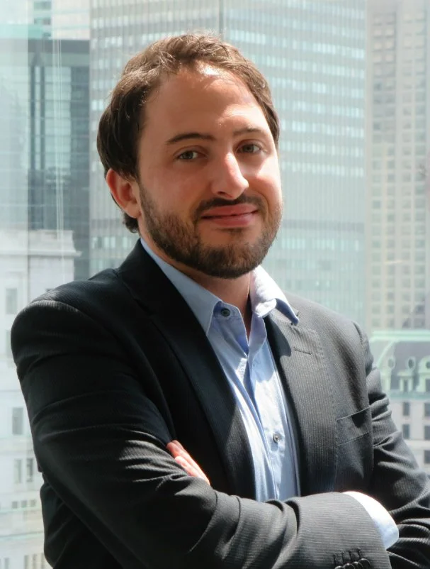 A man with dark brown hair and a beard, wearing a dark suit jacket and light blue shirt, standing with arms crossed in front of a modern glass building.