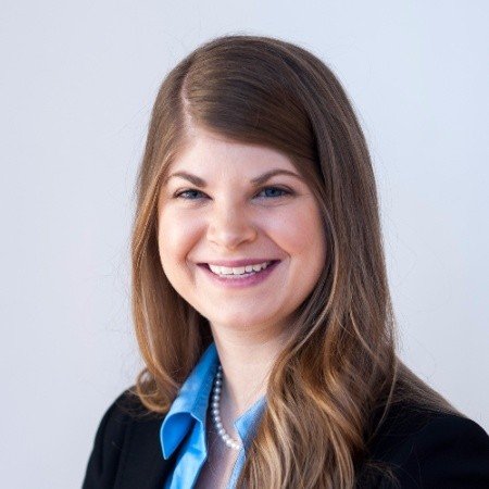 A woman with long brown hair smiling, wearing a black blazer over a blue shirt, with a pearl necklace.
