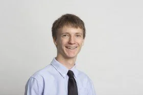 A young man smiling, wearing a light blue dress shirt and dark tie, standing against a plain light background.