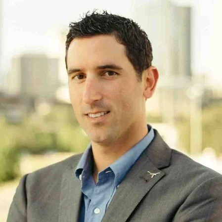 A man wearing a gray blazer and blue shirt standing outdoors in a cityscape background.