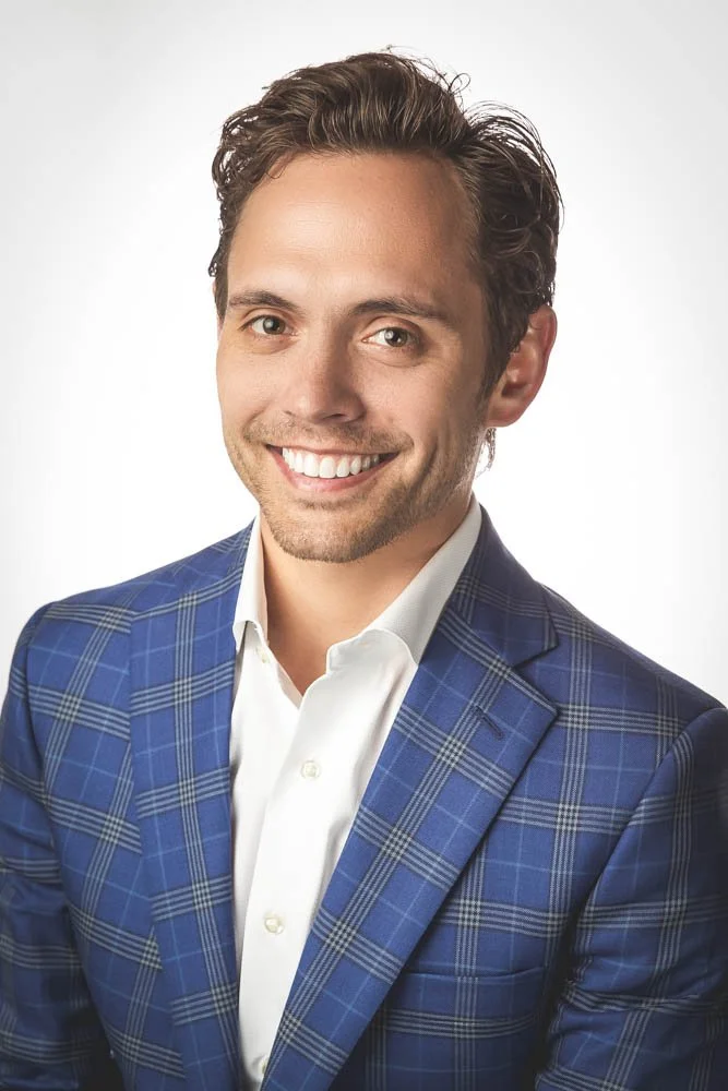 Young man with dark brown hair, smiling, wearing a white shirt and a blue plaid suit jacket, against a plain light background.
