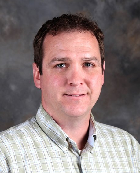 Portrait of a man in a light green plaid shirt against a gray mottled background.