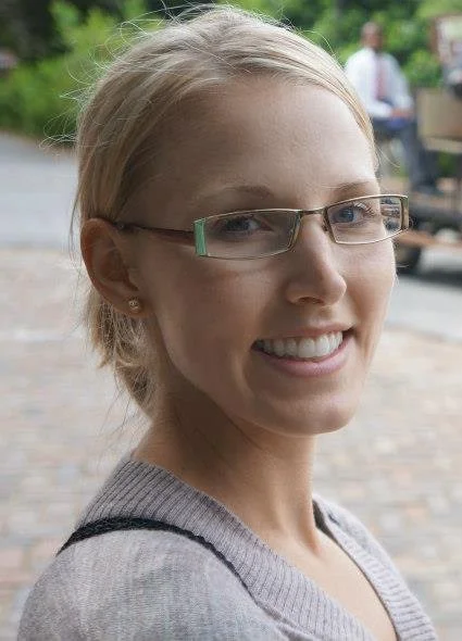 A woman with blonde hair, glasses, and a gray sweater smiling outdoors with blurred trees and people in the background.