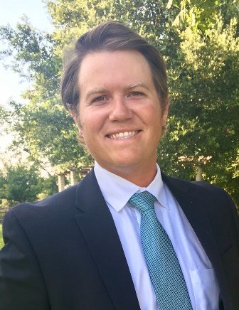 A young man in a suit smiling outdoors with trees in the background.