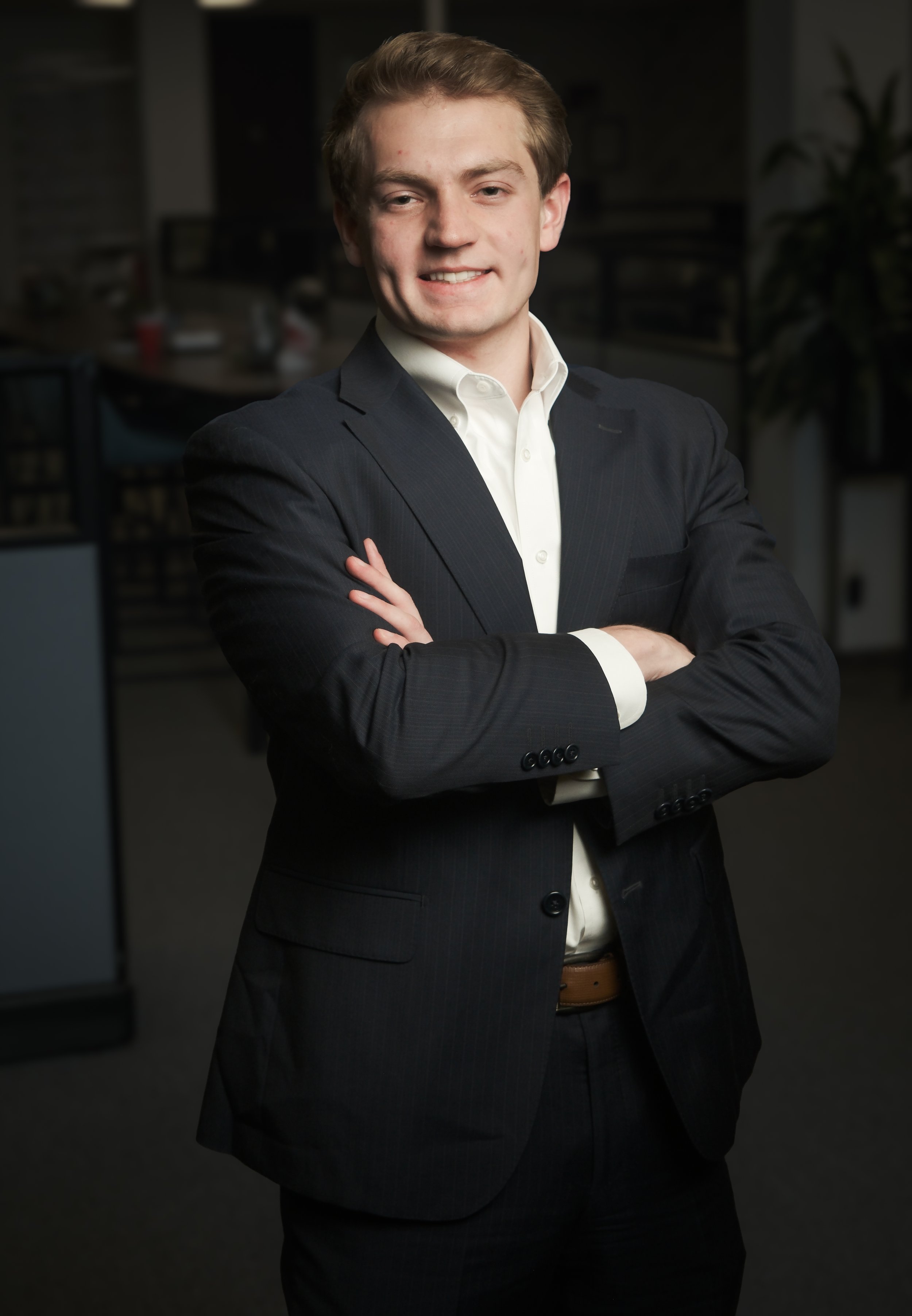 Young man with blonde hair smiling, wearing a black polo shirt and a red stole, standing outdoors against a beige wall.