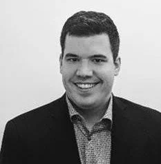A young man with short dark hair, smiling, wearing a dark blazer over a patterned shirt