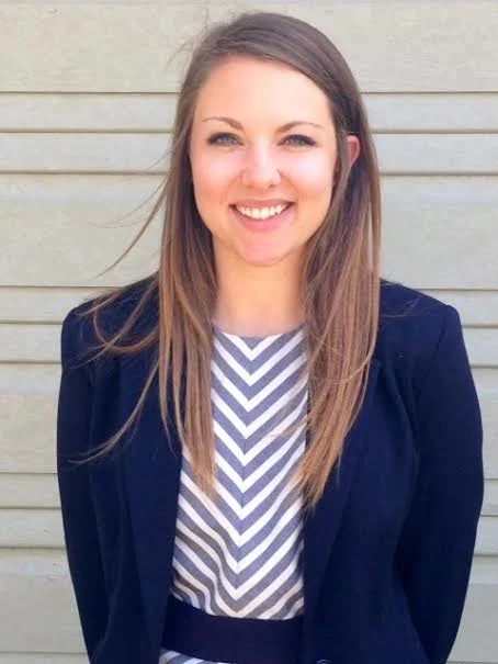 A young woman with long brown hair smiling, wearing a navy blazer over a striped top, standing in front of horizontal wooden siding.
