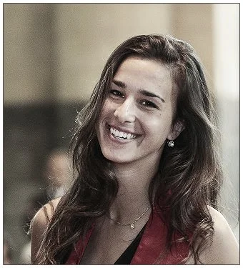 A young woman with long brown hair smiling at the camera in an indoor setting.