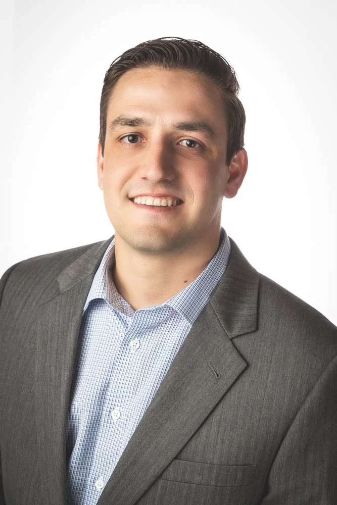 Portrait of a young man in a gray suit and light blue checkered shirt smiling at the camera against a white background.