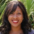 A woman with long, wavy dark hair smiling outdoors with green foliage in the background.