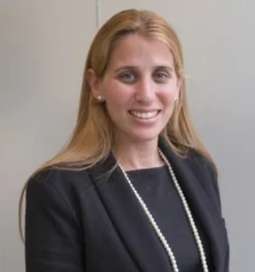 Portrait of a smiling woman with blonde hair wearing a black blazer and pearl necklace, standing against a plain background.