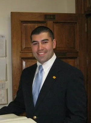 A man in a black suit and light blue tie, smiling, standing indoors in front of a wooden door.
