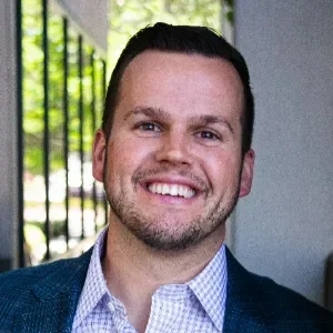 Man with dark hair and a beard smiling, wearing a blue blazer and light blue shirt, standing outdoors with greenery and a black iron fence in the background.
